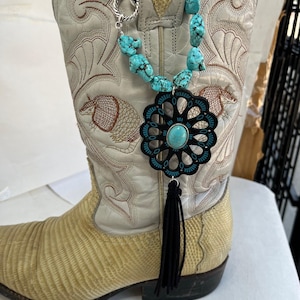 May include: A turquoise stone necklace with a black floral pendant and black tassel hangs from a white cowboy boot. The boot has embroidered details and a textured, light yellow leather shaft.