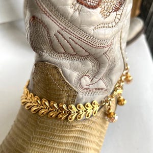 May include: A gold-colored anklet with a leaf design and dangling charms adorns a cream-colored cowboy boot. The boot features intricate stitching and a textured, light brown leather foot.