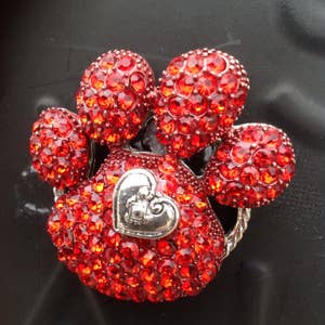Peut inclure: Broche en forme de patte avec un motif de cœur. La broche est recouverte de petits cristaux rouges scintillants. Un cœur argenté est au centre de l'empreinte de patte. La broche a une bordure en métal argenté.