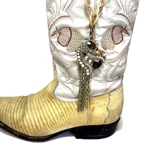 May include: A white cowboy boot with intricate brown embroidery and a textured, light yellow foot. A gold-toned chain holds a pendant with brown and clear gemstones and dangling chains. The boot has a pointed toe and a black sole.