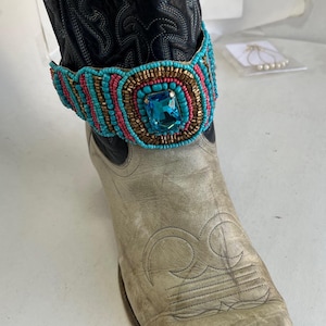 Op de afbeelding: Een met kralen versierde laarscuff met een grote vierkante blauwe edelsteen. De manchet heeft turquoise, roze en gouden kralen. De manchet zit op een versleten, gebroken witte cowboylaars met zwart leer boven de enkel.