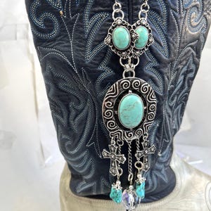May include: A silver-toned necklace with turquoise-colored stones and cross pendants hangs in front of a black and silver cowboy boot. The necklace features an oval turquoise stone in a decorative setting, with smaller stones and crosses dangling below.