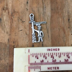 May include: Silver charm featuring a cartoon figure holding a street sign that reads "New Orleans". The figure is wearing a hat and holding a lamppost.