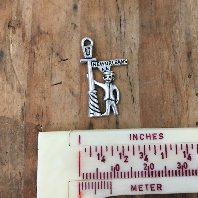 May include: Silver charm featuring a cartoon figure holding a street sign that reads "New Orleans". The figure is wearing a hat and holding a lamppost.