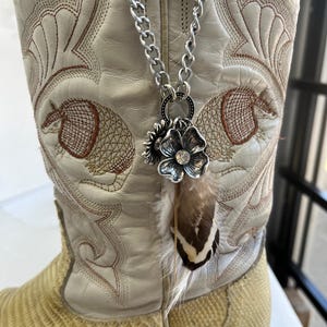 May include: A silver chain necklace with a flower pendant and a feather charm hangs in front of a cream-colored cowboy boot. The boot has intricate embroidery and a snakeskin-like texture on the lower part.
