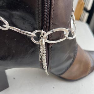 Könnte beinhalten: Eine silberne Stiefelkette mit Quasten-Detail ist an einem braunen Lederstiefel befestigt. Der Stiefel hat einen Reißverschluss und ein zweifarbiges Design mit einem dunkleren braunen Obermaterial und einer helleren braunen Spitze.