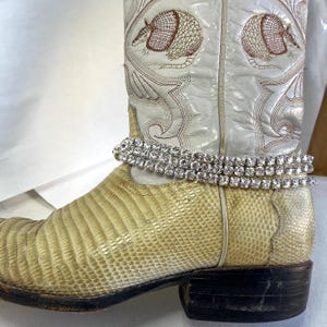May include: Cream-colored cowboy boot with lizard skin foot and white leather shaft. The shaft features embroidered armadillo designs. A double strand of sparkling rhinestones encircles the ankle area. The boot has a black sole and heel.