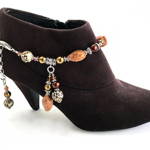May include: A brown suede ankle boot with a decorative silver chain and beaded anklet. The anklet features various beads in shades of brown, gold, and orange. The boot has a pointed toe and a high heel.