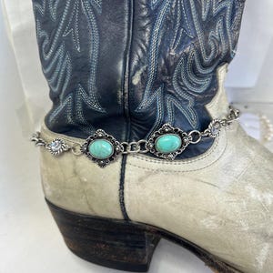May include: A silver-toned boot chain with turquoise-colored oval stones and clear crystals. The chain is draped over a worn, off-white and black cowboy boot with blue stitching.