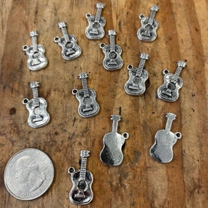 Pode incluir: Pingentes de guitarra prateados com uma moeda para comparação de tamanho. Os pingentes são detalhados com cordas, orifícios de som e pinos de afinação. Os pingentes estão espalhados em uma superfície de madeira.
