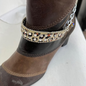 May include: A brown leather boot with a decorative leopard print ankle bracelet. The ankle bracelet is embellished with clear rhinestones and a silver chain with a small, iridescent charm. The boot has a pointed toe and a small heel.