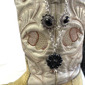 Op de afbeelding: Een zilveren ketting met zwarte stenen accenten hangt aan een crèmekleurige cowboylaars. De laars heeft ingewikkelde stiksels en een reliëf ontwerp. De ketting heeft een bloemvormige hanger en twee ovale stenen.