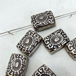 May include: Silver rectangular beads with a floral design. The beads have a raised, textured surface and are strung on a wire.
