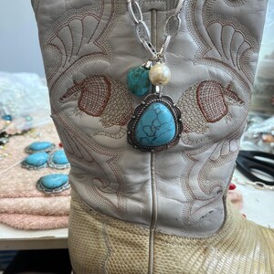 May include: A white leather cowboy boot with intricate embroidery and a silver chain necklace. The necklace features a large turquoise pendant, a pearl, and a smaller turquoise stone. The boot has a textured, light-colored lower section.