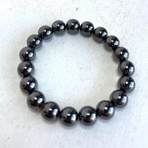 Op de afbeelding: Een donkergrijze kralenarmband. De armband is gemaakt van gladde, ronde kralen met een reflecterende, metallic glans. De kralen zijn aan elkaar geregen om een cirkelvorm te vormen. De armband wordt getoond op een wit oppervlak.