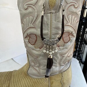 May include: A cream-coloured cowboy boot with detailed brown embroidery and a lizard-skin vamp. A decorative boot chain with a silver-toned filigree bar, faux pearls, and a brown and grey tassel hangs from the boot's pull straps.