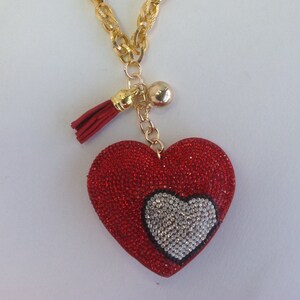 May include: A gold chain necklace with a red heart pendant. The heart is covered in sparkling red rhinestones and has a smaller white heart in the center. The necklace also has a small gold tassel and a gold ball charm.