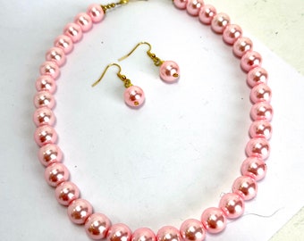 May include: A pink pearl necklace and matching earrings. The necklace is made of large, round pearls and has a gold clasp. The earrings are also made of large, round pearls and have gold hooks.