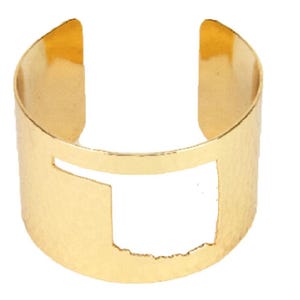 Può includere: Un bracciale a polsino color oro con un ritaglio dello stato dell'Oklahoma. Il bracciale ha una texture martellata e un design aperto, che consente di indossarlo e toglierlo facilmente. Questo accessorio è un gioiello elegante.