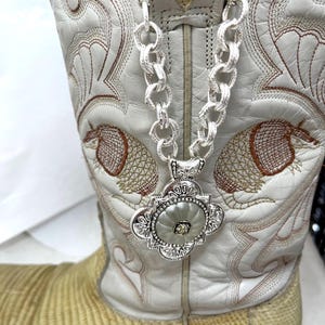 Puede incluir: Bota de vaquero de cuero blanco con costuras intrincadas y un collar de cadena plateada con un colgante decorativo. La bota tiene una textura similar a la piel de serpiente en la parte inferior.