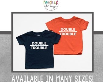 May include: Two children's t-shirts, one navy blue and one orange, each with the white text "DOUBLE TROUBLE" in a bold, sans-serif font. The shirts are displayed on a white background. The brand logo "heads up shirt designs" is at the top.