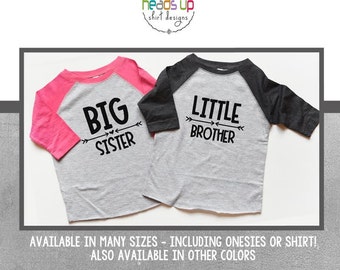 Puede incluir: Dos camisetas raglán para niños. Una es gris con mangas rosas y las palabras "BIG SISTER" en negro. La otra es gris con mangas gris oscuro y las palabras "LITTLE BROTHER" en negro. Las camisetas son de "heads up shirt designs".