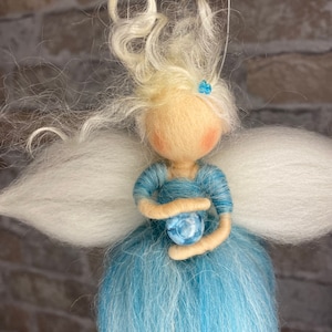 Könnte beinhalten: Eine Filz-Feenpuppe mit weißen Flügeln, blonden Haaren und einem blauen Kleid. Sie hält eine blaue Kugel in ihren Händen.