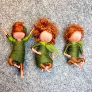 Könnte beinhalten: Drei handgefertigte Filzpuppen mit roten Garnhaaren, grünen Kleidern und braunen Armen und Beinen. Eine Puppe balanciert auf einem Bein, während die anderen sitzen. Die Puppen befinden sich auf einem grauen Filzhintergrund.
