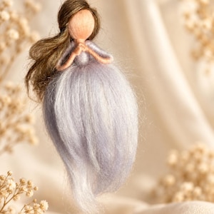 Könnte beinhalten: Ein hängendes Ornament in Form einer Figur mit langem braunem Haar und einem Kleid aus helllavendelfarbenen Fasern. Die Figur hat ein glattes, rundes, gesichtsloses Antlitz und die Arme vor der Brust verschränkt, aufgehängt an einem dünnen Faden.