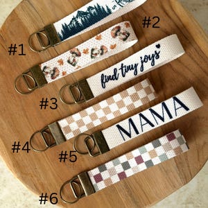 May include: Six fabric wristlet keychains with bronze-toned metal hardware. Designs include a mountain compass, mushrooms, the text "find tiny joys", a beige checkerboard, the text "MAMA", and a multi-colored checkerboard pattern.