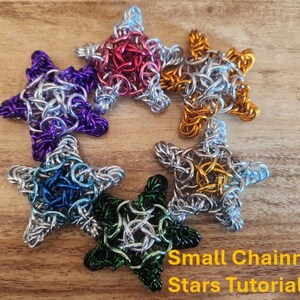 May include: Six small chainmail stars in different colors, including purple, blue, silver, green, gold, and red. The stars are arranged in a circle on a wooden surface. The text "Small Chainmail Stars Tutorial" is written below the stars.