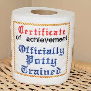 May include: White toilet paper roll with embroidered text reading "Certificate of achievement" and "Officially Potty Trained" in red and blue, framed with gold stitching. The roll is sitting on a woven surface.