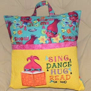 May include: A yellow and pink pillow with a pocket and a handle. The front of the pillow has an embroidered design of a troll with a book and the text "Sing & Dance & Hug & Read (a lot)". The back of the pillow has a blue and green fabric with a pattern of trolls and flowers.