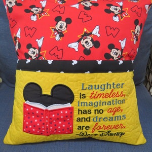 May include: A yellow pillow with a Mickey Mouse head and a red book with white stars. The pillow has a black and white Mickey Mouse print pocket and a quote from Walt Disney: "Laughter is timeless, imagination has no age, and dreams are forever."