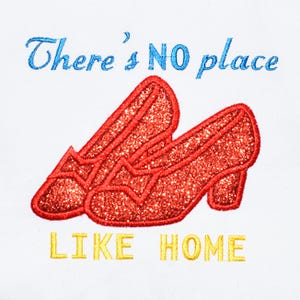 May include: An embroidered design featuring two red glittery shoes positioned diagonally. Above the shoes, the text reads "There's NO place" in blue, and below the shoes, the text reads "LIKE HOME" in yellow, all set against a plain white background.