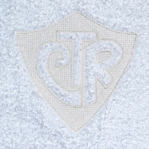 Puede incluir: Primer plano de un textil azul claro con un escudo bordado en blanco. El escudo presenta las letras "CTR" dentro de un borde en forma de escudo. El fondo tiene un aspecto texturizado y moteado.