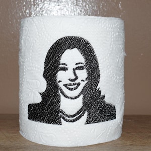 May include: A white toilet paper roll with a black and white embroidered image of a woman with long hair.