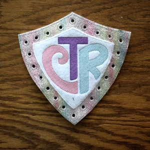 May include: A white felt shield with a pink "C", a purple "T", and a blue "R" embroidered on it. The shield has a silver glitter border with holes around the edge.