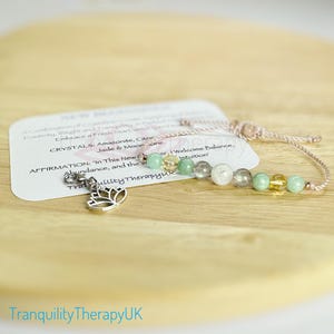 May include: A bracelet with a lotus charm and a variety of green, yellow, and grey stones. The bracelet is made with a light brown cord and has the text "In This New Chapter, I Welcome Balance, Intuition" on a card.