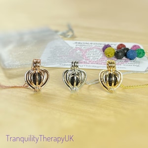 May include: Three heart-shaped aromatherapy diffuser necklaces in rose gold, silver, and gold. Each pendant holds a dark gray lava bead and hangs on a delicate chain. A small bag and a card with text are in the background.