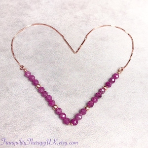 May include: A delicate rose gold chain necklace with a pink gemstone pendant. The chain is adjustable and features a slider clasp. The pendant is a round, faceted gemstone with a deep pink hue.