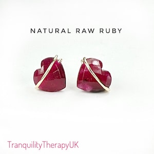 以下が含まれることがあります： 金色のワイヤーのアクセントが付いたハート型の原石ルビーのイヤリングのペア。イヤリングは白い背景に置かれています。NATURAL RAW RUBYはイヤリングの上に書かれています。TranquilityTherapyUKはイヤリングの下に書かれています。