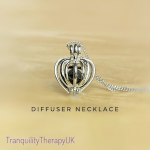 May include: Silver heart-shaped diffuser necklace with a black lava stone bead inside. The necklace is on a wooden surface. DIFFUSER NECKLACE and TranquilityTherapyUK are written below the necklace.