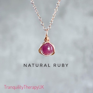 May include: A delicate rose gold chain necklace featuring a natural ruby gemstone wrapped in wire. The pendant is a single round ruby with a deep pink hue. The text "NATURAL RUBY" is displayed below the pendant.
