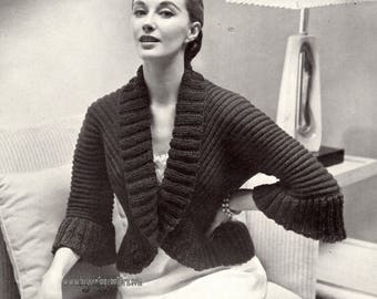 May include: A black knitted cropped cardigan with a wide collar and long sleeves. The cardigan is worn by a woman with dark hair and a white dress.