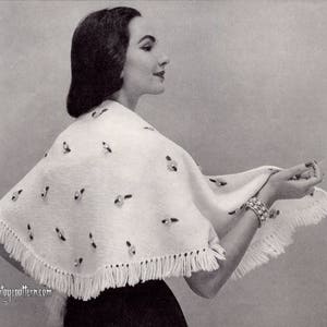 May include: A white knitted shawl with a fringe border and small white embellishments. The shawl is draped over a woman's shoulders.