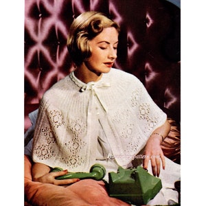 Peut inclure: Une cape en tricot blanc avec un motif en dentelle. La cape a un col et une cravate au cou. Le mannequin porte la cape par-dessus une robe blanche et est assis sur un canapé rose capitonné.