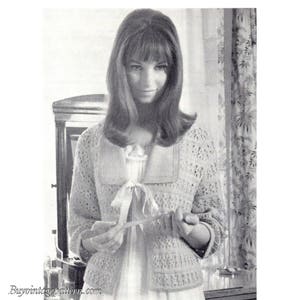 May include: A black and white photo of a woman wearing a crocheted cardigan with a lace pattern. The cardigan has a collar and a bow at the front. The woman is holding a small object in her hands.