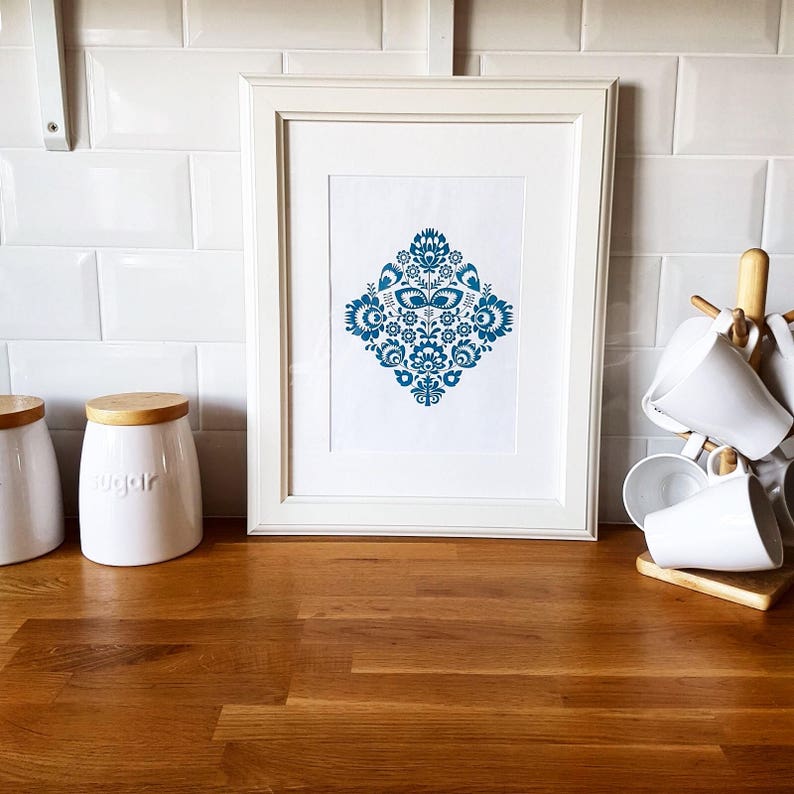 Peut inclure: Un cadre blanc avec une impression de motif d'art populaire bleu et blanc. L'impression est en forme de diamant avec des motifs floraux. Le cadre est pos&eacute; sur un comptoir en bois.