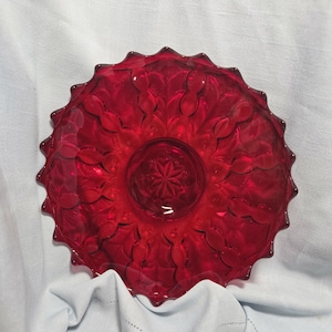 May include: A circular ruby red glass bowl featuring a scalloped edge and a pressed pattern of repeating oval shapes radiating from a central starburst design. The glass is translucent and deep red, showcasing a detailed geometric texture.